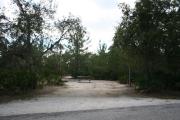 Photo is at the front of LOOP 1 #4 looking out towards back of site; gray campground road with green trees surrounding the site, also on the back of the site is 2 clothes line poles,  to the far right of site is water and electric hookups; far left of site is circular ground grill and picnic table.