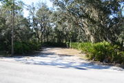 Photo is at the front of LOOP 1 #6 looking out towards back of site; gray campground road with green trees surrounding the site, also on the back of the site is 2 clothes line poles,  to the far right of site is water and electric hookups; far left of site is circular ground grill and picnic table.