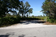 Photo is at the front of LOOP 1 #10 looking out towards back of site; gray campground road with green trees surrounding the site, also on the back of the site is 2 clothes line poles,  to the far right of site is water and electric hookups; far left of site is circular ground grill and picnic table. Also in the distance you have a lake view
