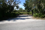 Photo is at the front of LOOP 1 #23 looking out towards back of site; gray campground road with green trees surrounding the site, also on the back of the site is 2 clothes line poles,  to the far right of site is water and electric hookups; far left of site is circular ground grill and picnic table.