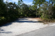 Photo is at the front of LOOP 1 #25 looking out towards back of site; gray campground road with green trees surrounding the site, also on the back of the site is 2 clothes line poles,  to the far right of site is water and electric hookups; far left of site is circular ground grill and picnic table.