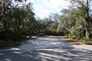 Photo is at the front of LOOP 1 #26 looking out towards back of site; gray campground road with green trees surrounding the site, also on the back of the site is 2 clothes line poles,  to the far right of site is water and electric hookups; far left of site is circular ground grill and picnic table.