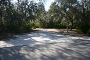 Photo is at the front of LOOP 1 #28 looking out towards back of site; gray campground road with green trees surrounding the site, also on the back of the site is 2 clothes line poles,  to the far right of site is water and electric hookups; far left of site is circular ground grill and picnic table.