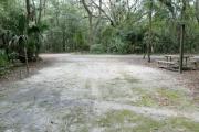 A September view of the campground drive from within campsite #7, the picnic table is seen on the right side of the tent/RV space, and the water hookup is seen on the left.  Another campsite is visible across the campground drive.  Site is surrounded on three sides by a mixed oak, pine, and cabbage palm forest.