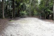 A September view of campsite #17 taken from the site entrance, with tent/RV space in center of image, grill and fire ring on left, picnic table in rear center of site, and water and electrical hookups on right hand side.  Site is surrounded on three sides by a mixed oak, pine, and cabbage palm forest.