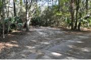A September view of campsite #18 taken from the site entrance, with tent/RV space in center of image, picnic table and fire ring on left hand side, and water and electrical hookups on right hand side.  Site is surrounded on three sides by a mixed oak, pine, and cabbage palm forest.