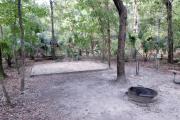 A September view of tent site #19, showing raised tent pad, with fire ring in foreground, water and electrical hookups to the right, and picnic table on right edge of picture. Site is surrounded on three sides by a mixed oak, pine, and cabbage palm forest.