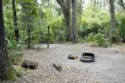 A September view of tent site #26, showing raised tent pad, fire ring, and lantern post.  Picnic table, water and electrical hookups are a short distance away.  Site is surrounded on three sides by a mixed oak, pine, and cabbage palm forest.