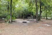 A September view of tent site #28, showing raised tent pad, fire ring, picnic table and lantern post.  Water and electrical hookups are a short distance away.  Site is surrounded on three sides by a mixed oak, pine, and cabbage palm forest.  Campground loop drive is visible through woods on right.