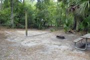 A September view of tent site #29, showing raised tent pad, fire ring, picnic table and lantern post.  Water and electrical hookups are a short distance away.  Site is surrounded on three sides by a mixed oak, pine, and cabbage palm forest. Campground loop drive is visible through woods on right.