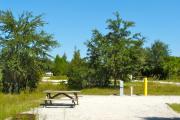 A gravel sitting area with a wooden picnic table, a cast-iron fire circle, water, and electric hookups. A couple of lower trees and bushes are between the sites.