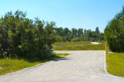 A 55 feet long gravel back up campsite with trees to the sides and a field of low growing vegetation in the background. The road leading into the campground can be seen in the background. The sitting area is located to the left and has a wooden picnic table and a cast-iron fire circle.