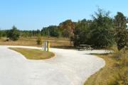 A 60 feet long gravel pull through campsite with vegetation giving privacy to the left side of the site. The sitting area is also located to the left and has a wooden picnic table and a cast-iron fire circle. Water and electric hook-ups are on the left hand side of the site.