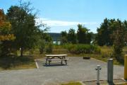 Water and electric hookups in the foreground across from the sitting area that has a wooden picnic table and a cast-iron fire circle. Hammond Lake is visible in the background.