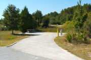 A 60 feet long gravel back up campsite with some trees and low growing vegetation around the site. The sitting area is located to the left and has a wooden picnic table and a cast-iron fire circle. Water and electric hook-ups are on the right hand side of the site.
