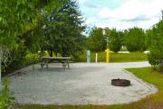 A gravel sitting area with a cast-iron fire circle and a wooden picnic table with vegetation in the background.