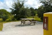 A gravel sitting area with a wooden picnic table and a cast-iron fire circle. Some trees and bushes are giving partial shade and some privacy.