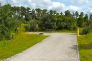 A 45 feet long gravel back up campsite with some low growing vegetation in the background. The sitting area is located to the left and has a wooden picnic table and a cast-iron fire circle. Water and electric hook-ups are on the right hand side of the site.