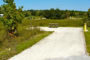 A 45 feet long gravel back up campsite with low growing vegetation around it. The sitting area is located to the left and has a wooden picnic table and a cast-iron fire circle. Water and electric hook-ups are on the right hand side of the site.