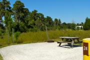 A gravel sitting area with a wooden picnic table, a cast-iron fire circle, and a wooded backdrop.
