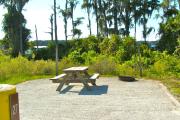 A gravel sitting area with a wooden picnic table, a cast-iron fire circle, and a wooded backdrop. Dixie Lake can be seen behind the cypress trees.