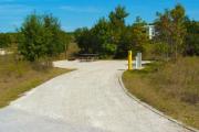 A 50 feet long gravel back up campsite with low growing trees and vegetation around it. The sitting area is located to the left and has a wooden picnic table and a cast-iron fire circle. Water and electric hook-ups are on the right hand side of the site.