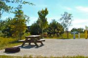 A gravel sitting area with a wooden picnic table and a cast-iron fire circle. A few trees and bushes are giving some privacy between the sites.