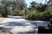 Looking out of site toward campground road; picnic table and standing grill on left; water spigot and electrical hook up on right; site is paved and a sidewalk leads to the bath house