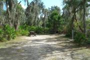 Looking into campsite from campground road; water spigot and electrical hook up on right; picnic table and fire ring are in rear of site; site is mixture of grass and sand