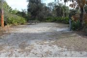 Looking into site from campground road; site mostly grass and sand; water spigot and electrical hook up on right; picnic table and fire ring at rear of site