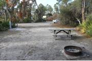 Looking out of site toward campground road; site is mostly grass and sand; water spigot and electrical hook up on left; fire ring and picnic table at rear of site