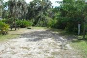 Looking into site from campground road; water spigot and electrical hook up on right; picnic table and fire ring on left; site mixture of grass and sand