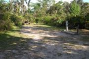 Looking into site from campground road; water spigot and electrical hook up on right; picnic table and fire ring on left; site mixture of grass and sand, mostly shaded in afternoon