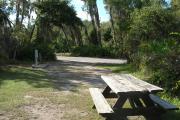 Looking out of site toward campground road; fire ring and picnic table on right; water spigot and electrical on left; site mixture of grass and sand, partially shaded