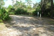 Looking into site from campground road; water spigot and electrical hook up on right; picnic table and fire ring at rear of site; site mixture of grass and sand