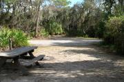 Looking out of site toward campground road; fire ring and picnic table on left; water spigot and electrical on left; site mixture of grass and sand, partially shaded