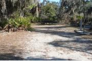 Looking into site from campground road; water spigot and electrical hook up on right; picnic table and fire ring at rear of site; site mixture of grass and sand