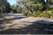 Looking out of site toward campground road; fire ring and picnic table on right; water spigot and electrical on left; site mixture of grass and sand, partially shaded