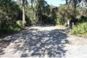 Looking into site from campground road; water spigot and electrical hook up on right; picnic table and fire ring on right; site mixture of grass and sand