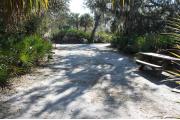 Looking out of site toward campground road; fire ring and picnic table on right; water spigot and electrical on left; site mixture of grass and sand, partially shaded