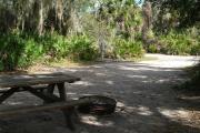 Looking out of site toward campground road; fire ring and picnic table on right; water spigot and electrical on left; site mixture of grass and sand, partially shaded