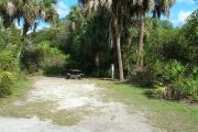 Looking into site from campground road; water spigot and electrical hook up on right; picnic table and fire ring on right; site mixture of grass and sand