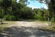 Looking into site from campground road; water spigot and electrical hook up on right; picnic table and fire ring on left; site mixture of grass and sand