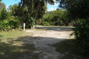 Looking out of site toward campground road; fire ring and picnic table on right; water spigot and electrical on left; site mixture of grass and sand, partially shaded