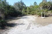 Looking into site from campground road; water spigot and electrical hook up on right; picnic table and fire ring on left; site mixture of grass and sand