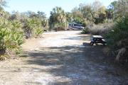 Looking out of site toward campground road; fire ring and picnic table on right; water spigot and electrical on left; site mixture of grass and sand, partially shaded