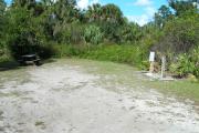 Looking into site from campground road; water spigot and electrical hook up on right; picnic table and fire ring on left; site mixture of grass and sand