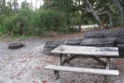 View of a wooden retaining wall, picnic table and fire ring in shade.