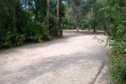 View of a vacant campsite with a fire ring/grill and picnic table. The main part of the site is sandy with sparse vegetation and a mix of trees to the sides. There is partial shade.