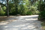 View of a vacant campsite with a picnic table, a fire ring/grill, electric pedestal, and water spigot. There is a large oak tree which provides partial shade.